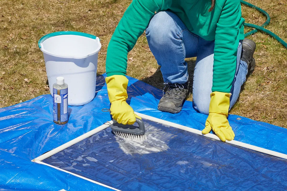 Woman wearing rubber gloves scrubs window screens lying on a blue tarp.